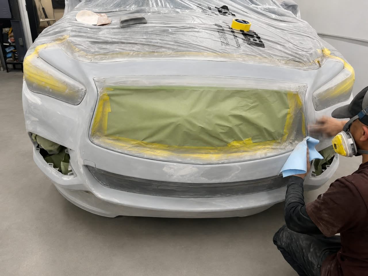 Technician in a respirator preps the front bumper of a white Infiniti for paint — Total Care Collision Pompano Beach FL
