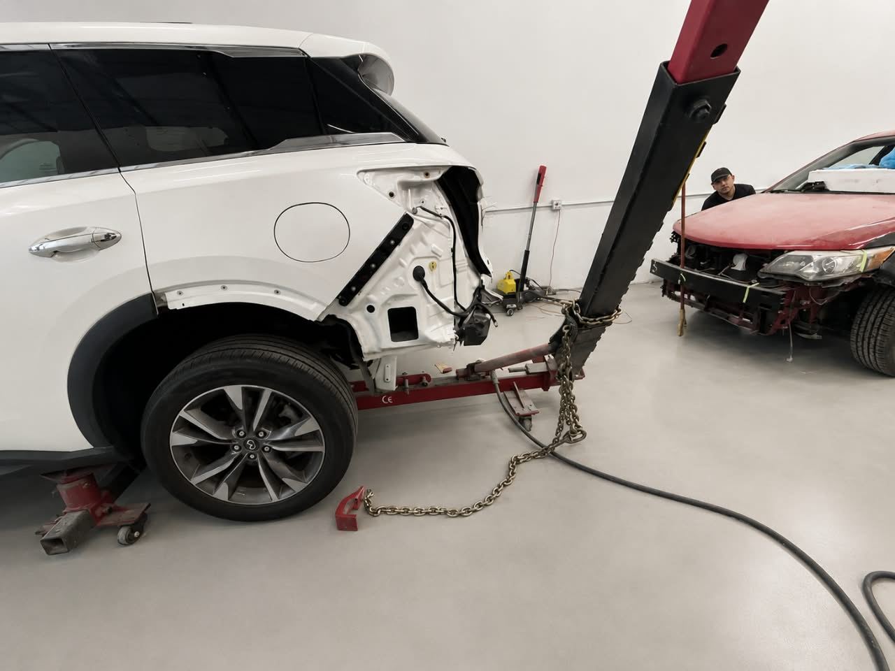 White Infiniti chained to the frame-straightening machine, technician at work — Total Care Collision Pompano Beach FL