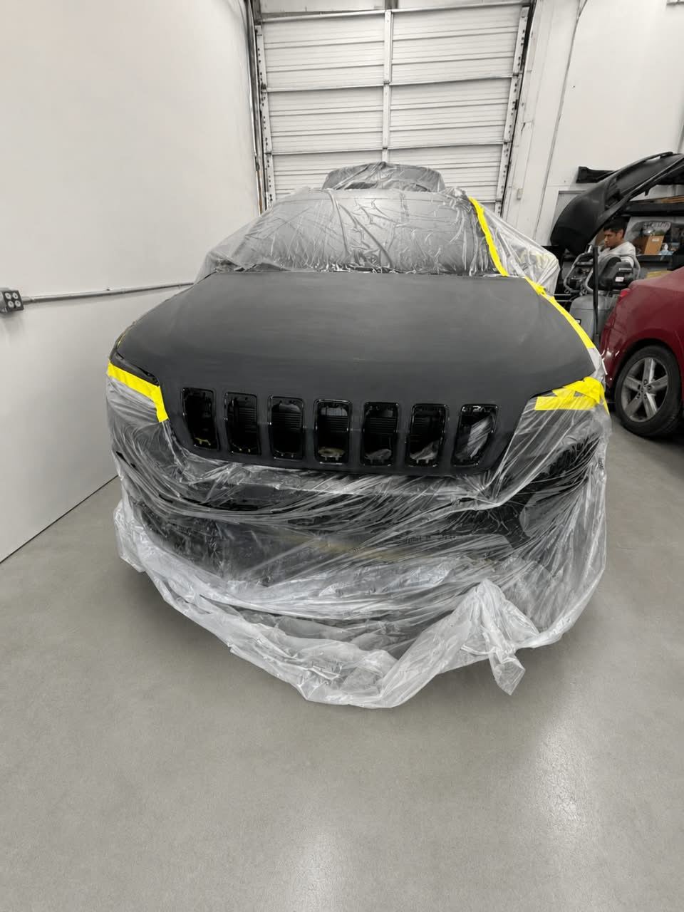 Jeep Cherokee masked in plastic with a primed black hood, technician in the background — Total Care Collision Pompano Beach FL