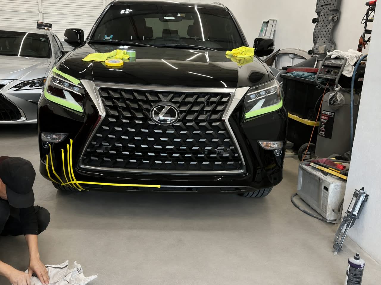 Technician kneeling to tape the front bumper of a black Lexus GX before paint — Total Care Collision Pompano Beach FL
